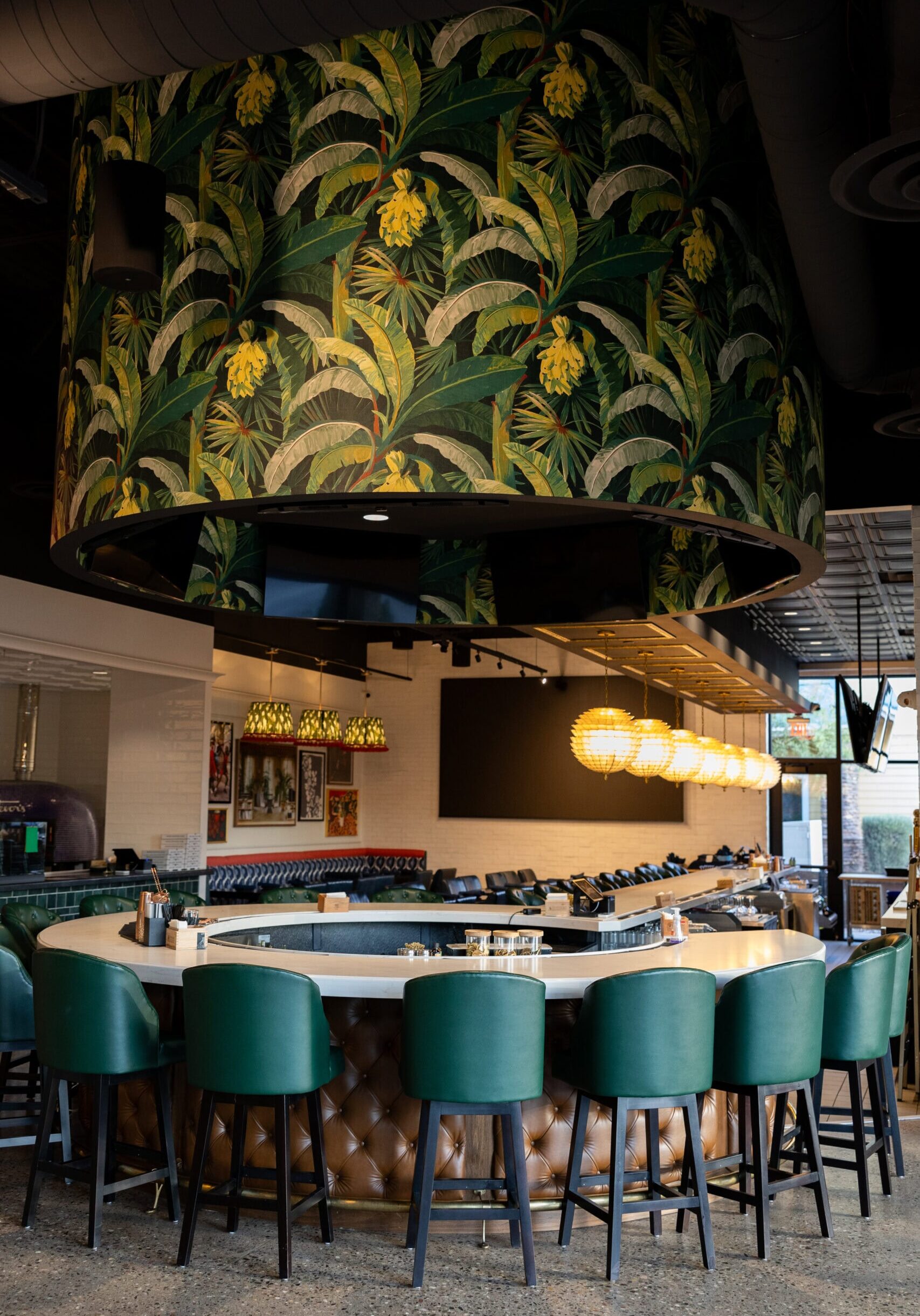 A modern bar with green upholstered chairs, a round counter, and a large overhead structure decorated with tropical leaf wallpaper. Spherical pendant lights hang in the background.