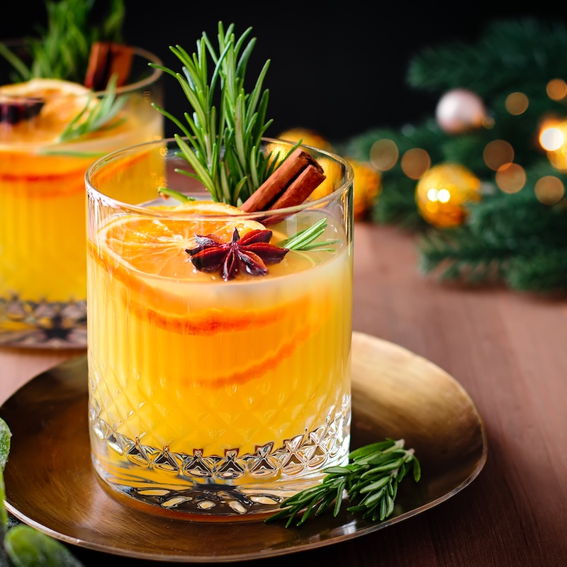 Two glasses of orange cocktail garnished with orange slices, rosemary, star anise, and cinnamon sticks sit on a gold tray by Crimson Design Group, surrounded by festive decorations in the background.