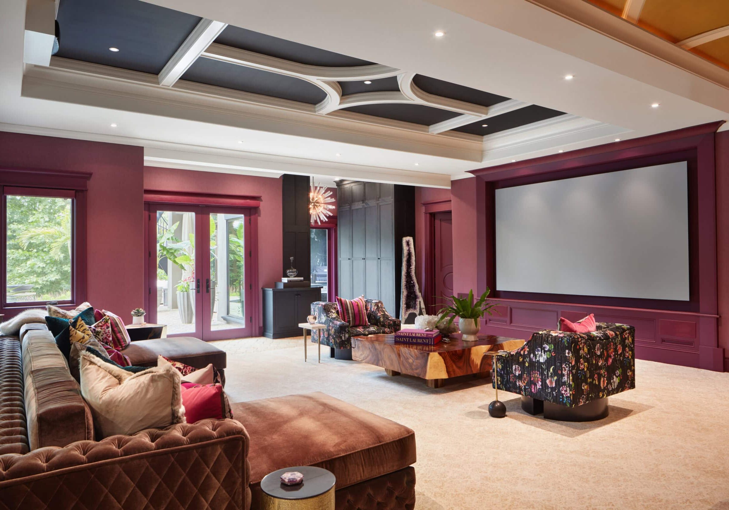 A spacious home theater room with a large projector screen, plush seating, decorative ceiling, and patterned furniture, featuring burgundy walls and natural light from windows.