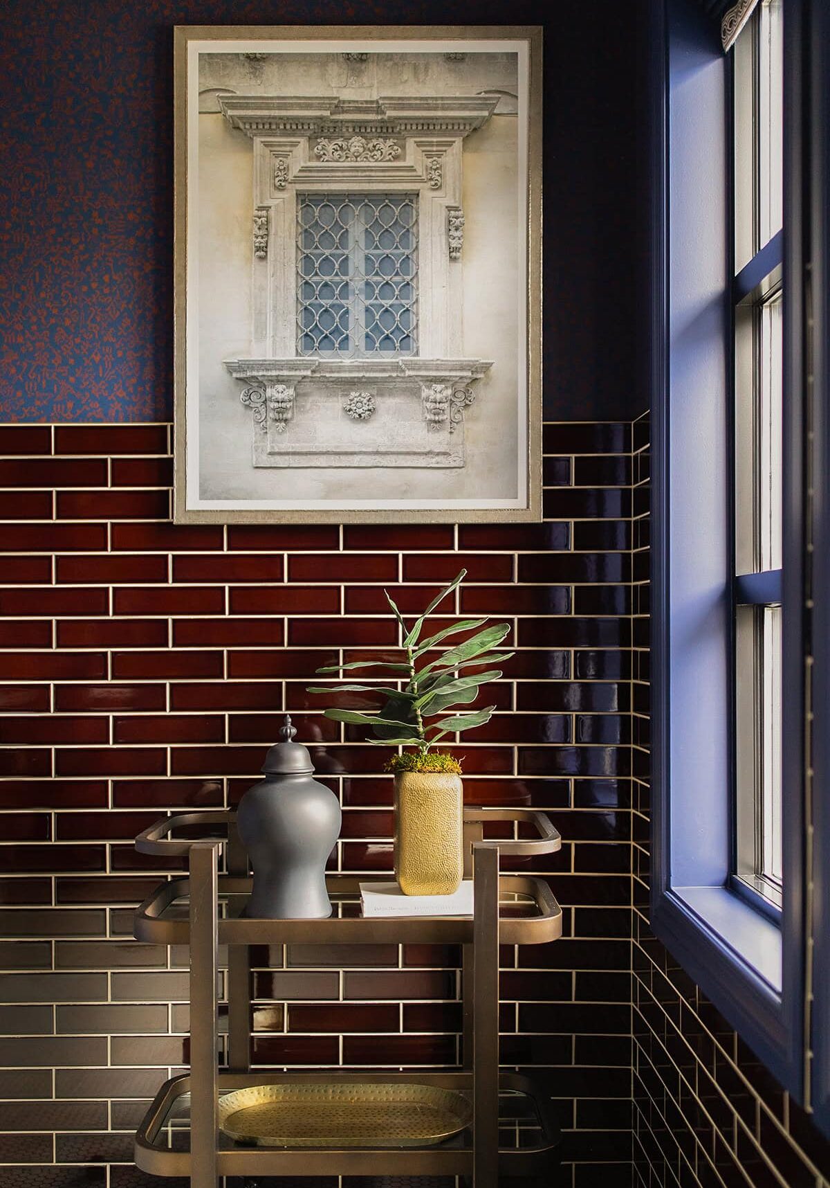A decorative bar cart with a dark vase, yellow planter, and plant stands against glossy brown tiles; above hangs framed art of an ornate window. Sunlight from the right highlights this Westerville Ohio-inspired nook.