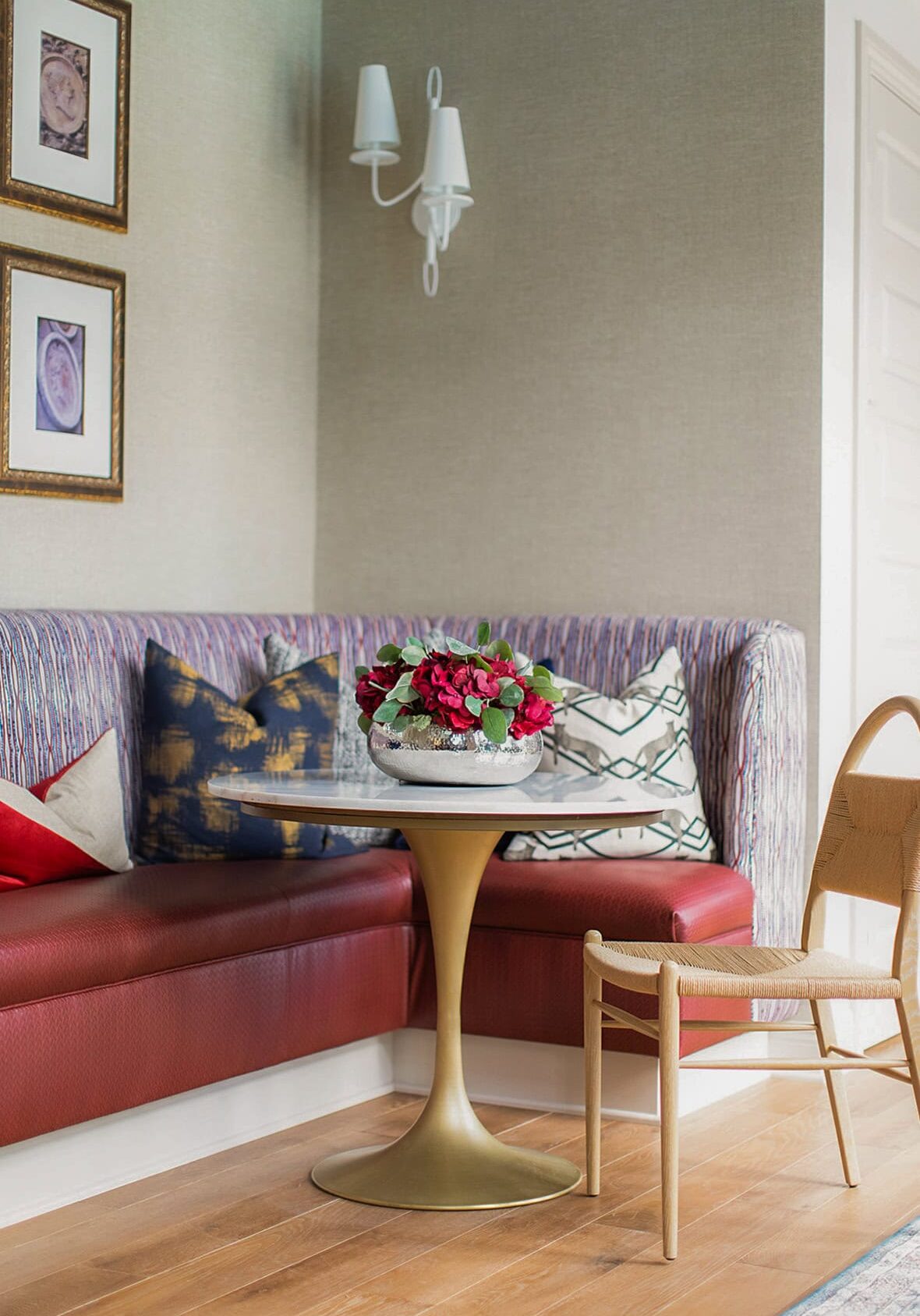 A small marble-top table with a flower arrangement sits by a red upholstered corner bench, patterned pillows, framed wall art, and a single wooden chair in a cozy Westerville Ohio room.