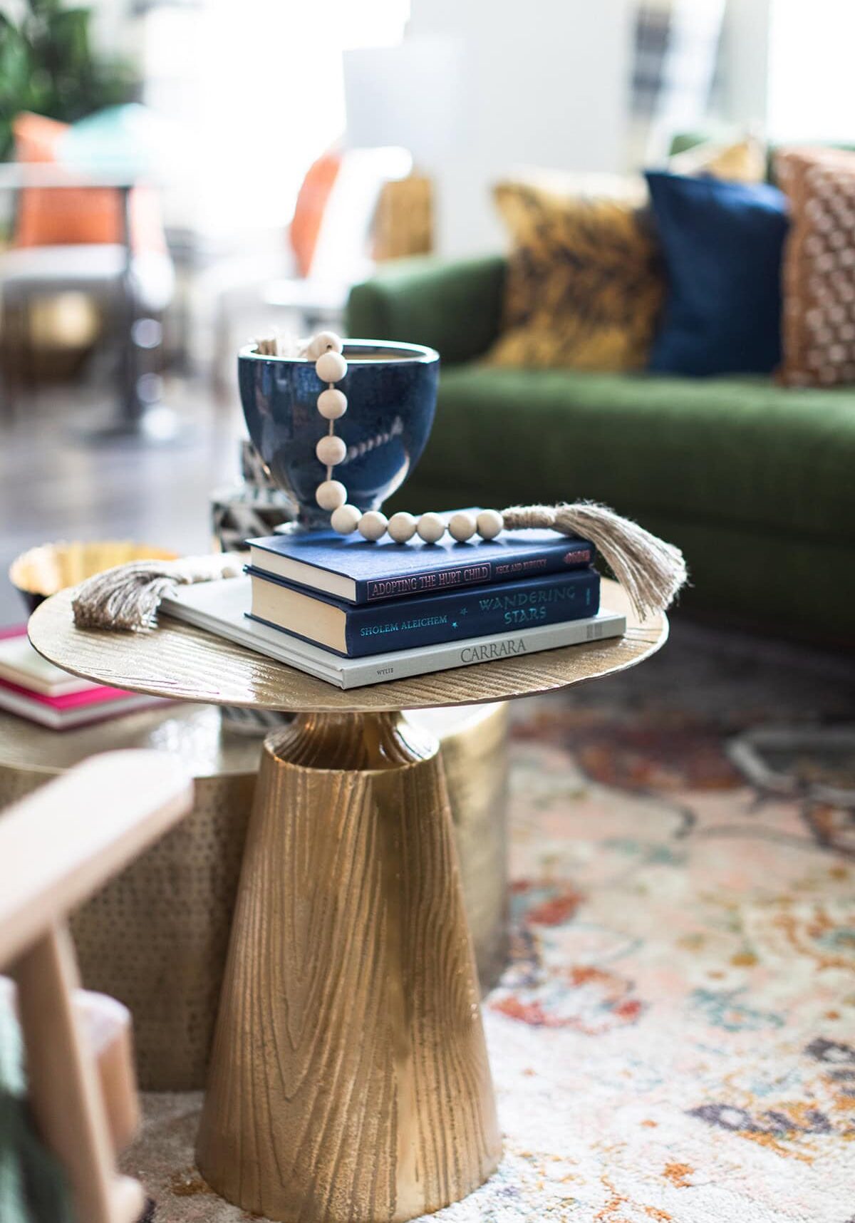 A small round gold side table holds stacked books, a blue bowl with wooden beads, and tassels in a bright, modern living room with a green sofa and patterned pillows.