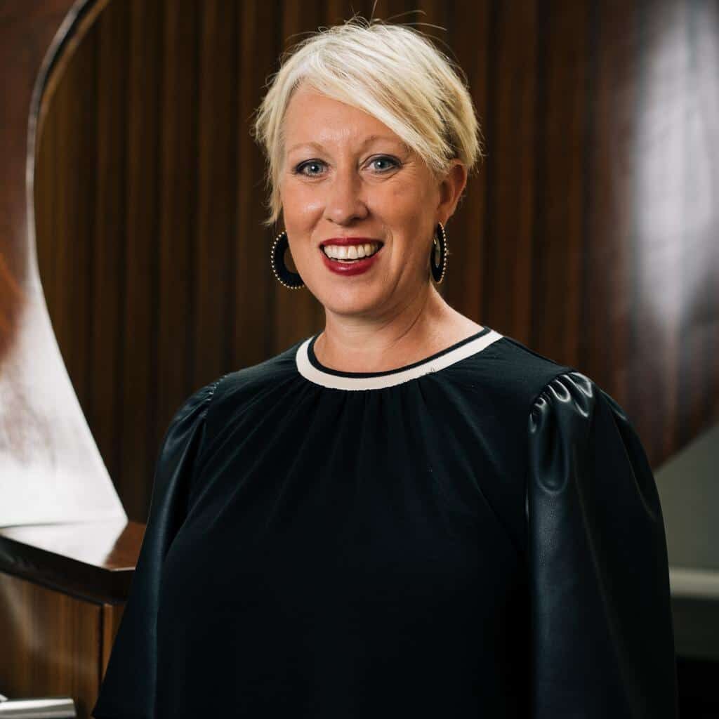 A woman with short blonde hair, wearing a black top with puffed sleeves, stands smiling in front of a wooden spiral staircase designed by Crimson Design Group.