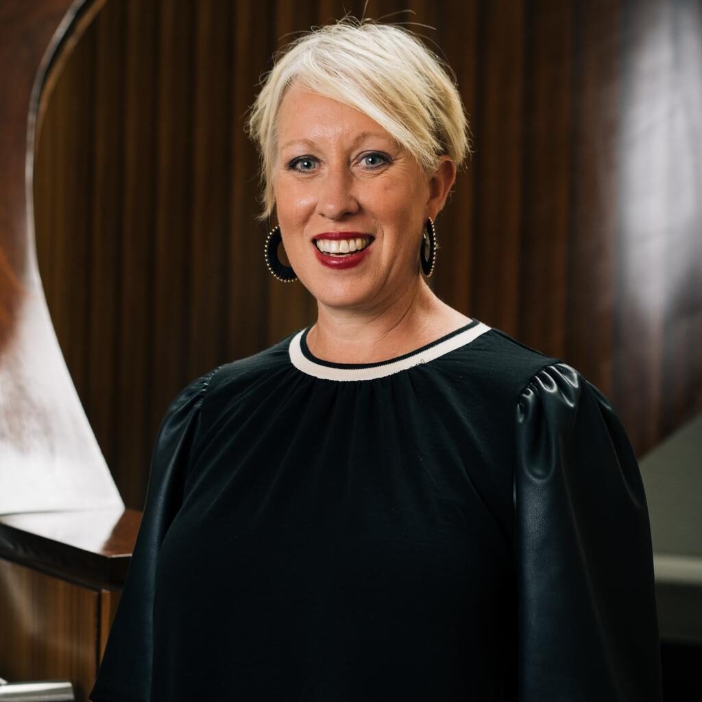 A woman with short blonde hair and hoop earrings, wearing a black dress with puff sleeves, stands in front of a wooden spiral staircase designed by Crimson Design Group.