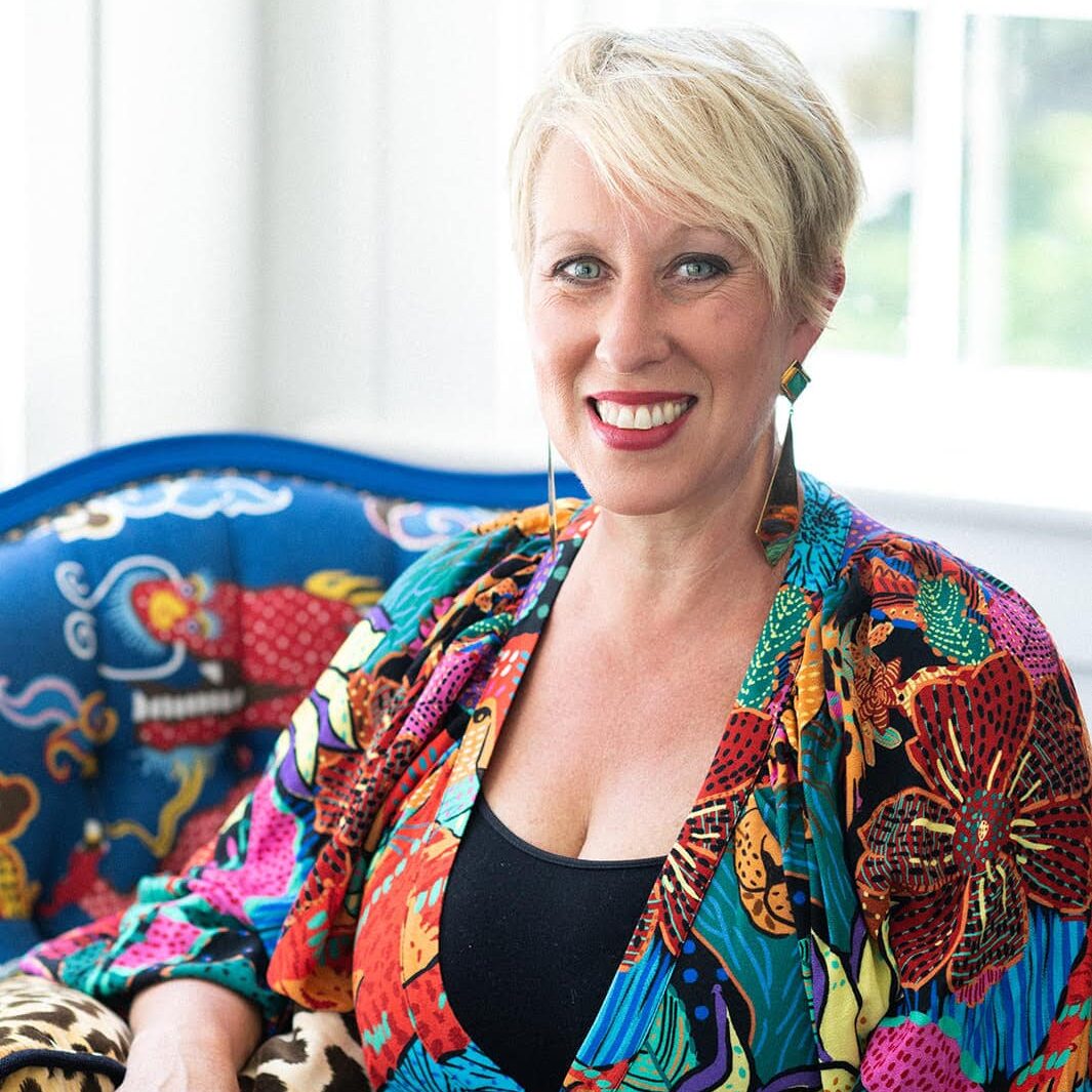 A woman with short blonde hair wearing a colorful, patterned outfit sits on a blue chair with a vibrant animal design by Crimson Design Group, smiling at the camera.