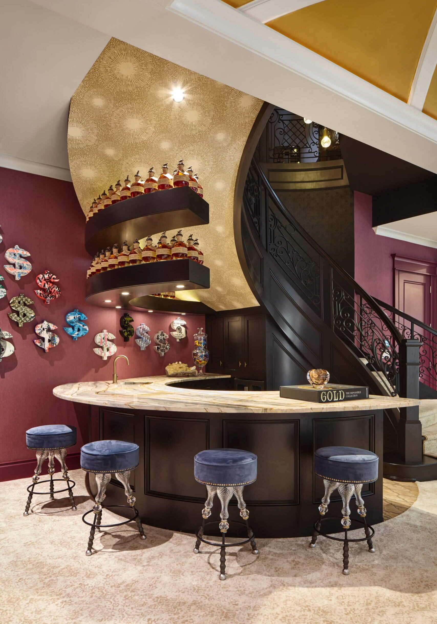 A curved staircase with ornate railing rises behind a modern bar with blue velvet stools, gold accents, and decorative shelves displaying colorful jars and art pieces on the walls.