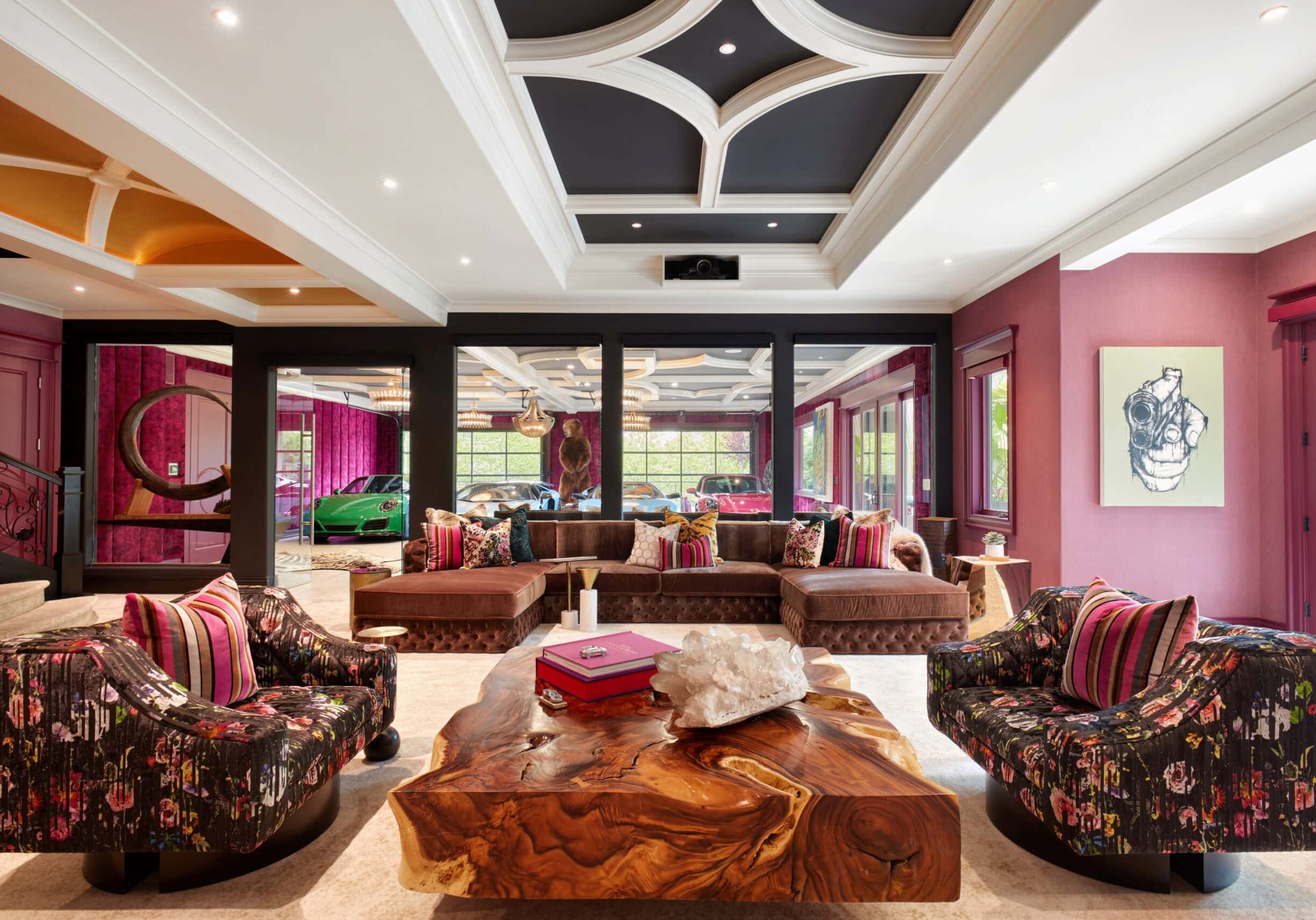 Spacious living room with patterned armchairs, a large wood coffee table, brown sofas, modern art on pink walls, and a view of cars through glass panels.