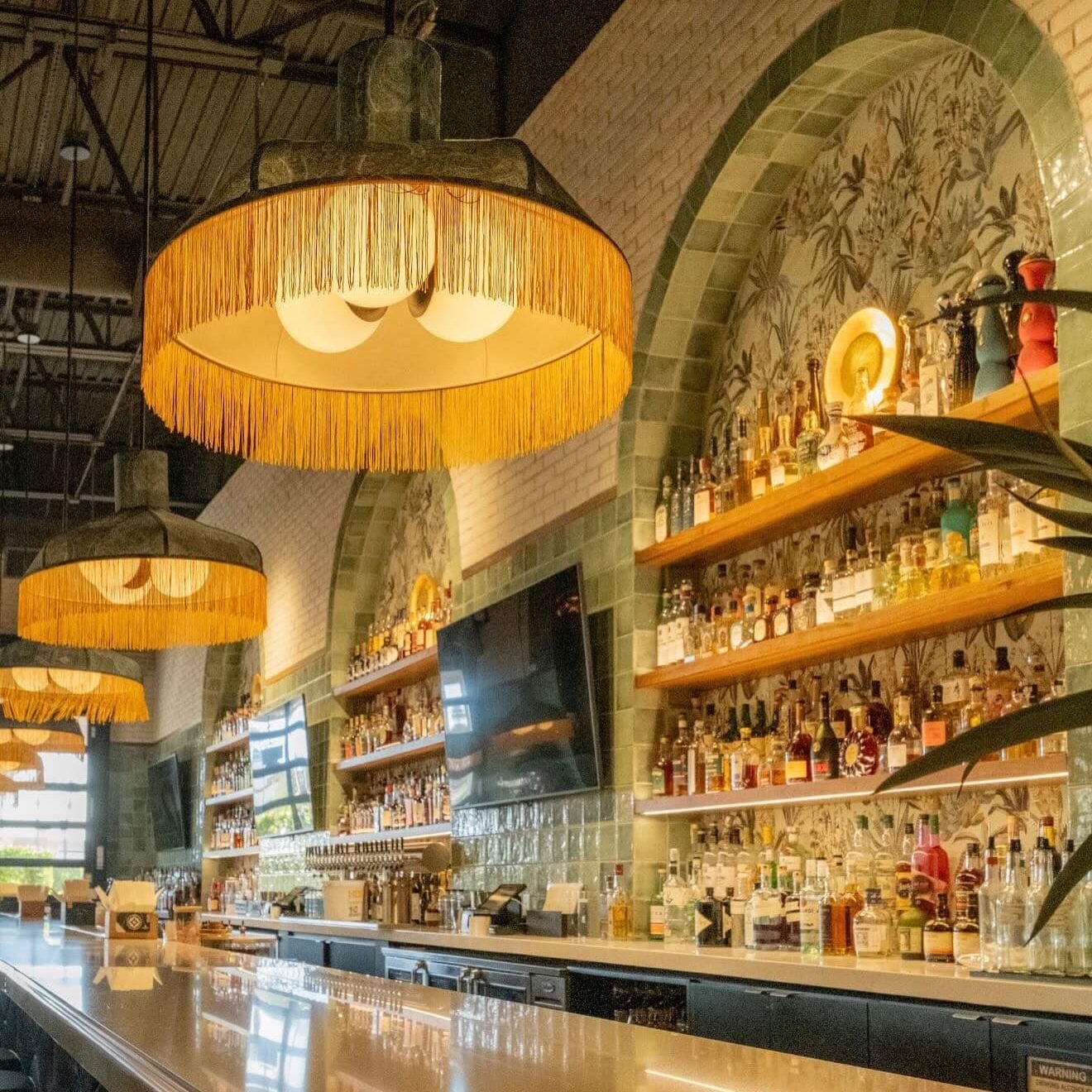 Contemporary bar interior with a long counter, hanging fringe lamps, shelves stocked with bottles, a wall-mounted TV, and decorative tiled arches.