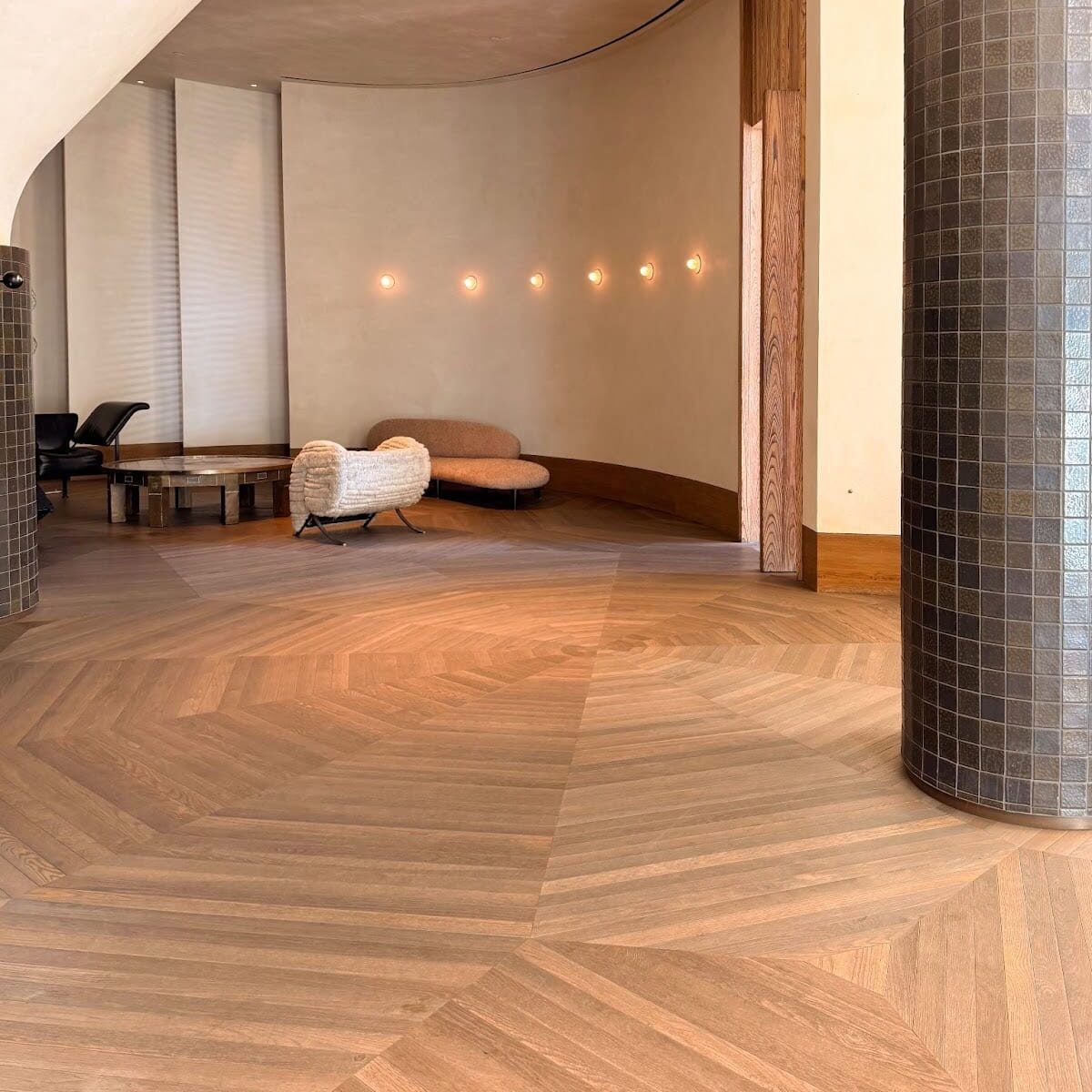 Modern interior with light wood chevron flooring, curved beige walls, tiled column, and minimalist furniture including a round coffee table and two lounge chairs.