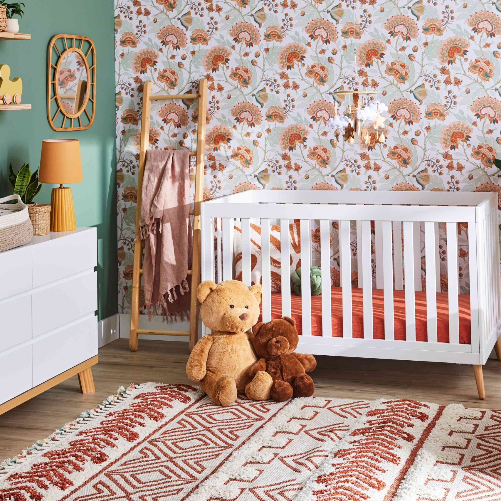 A nursery by Crimson Design Group with a white crib, two stuffed bears on a patterned rug, a dresser with changing pad, leafy plant, and floral wallpaper.