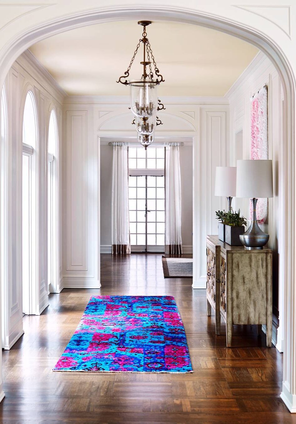 A bright entryway with white walls and arched windows features a bold blue and pink rug on dark wood floors, a chandelier, and a wooden console table with lamps.