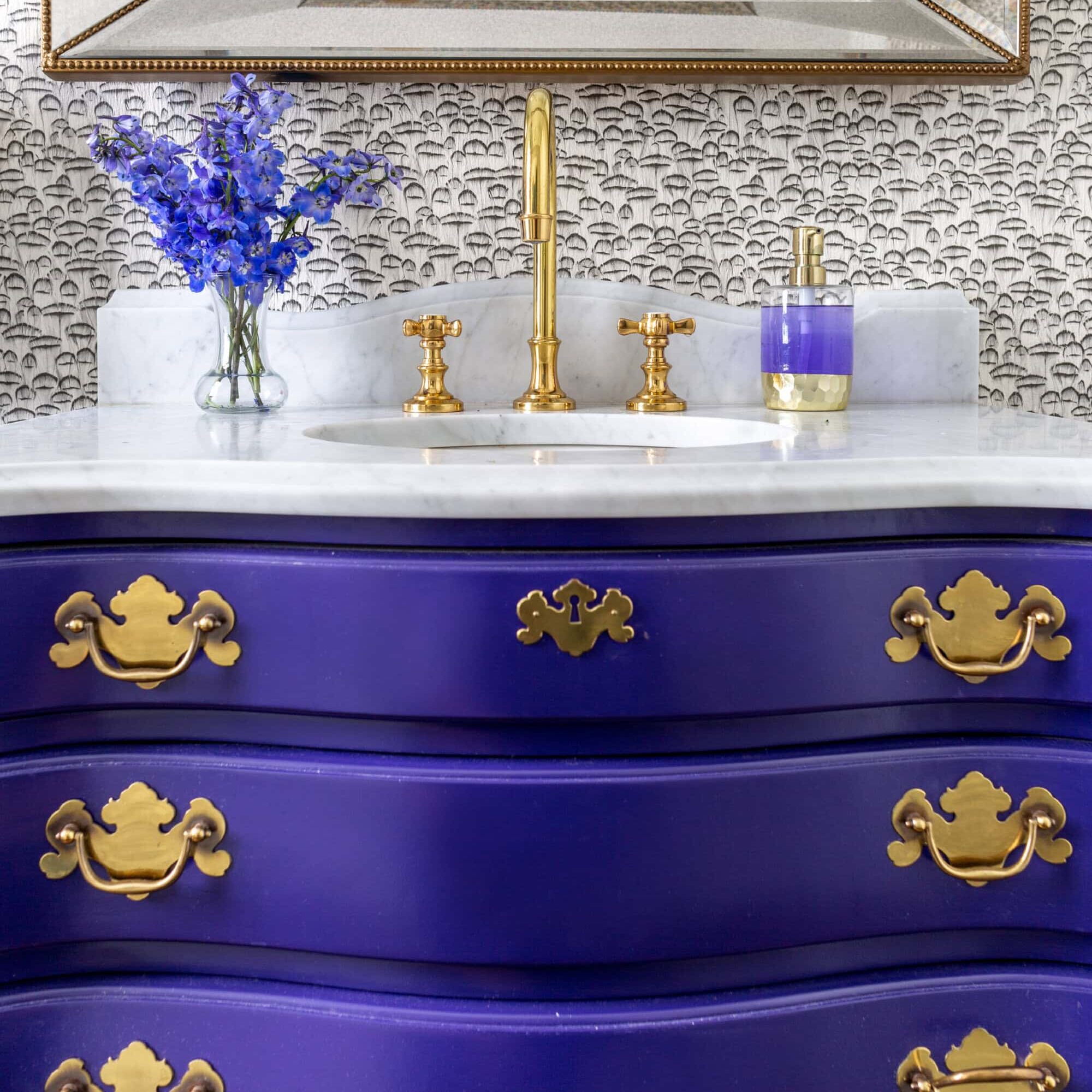 A bathroom vanity with a curved, deep purple cabinet, gold hardware, a marble countertop, gold faucet, purple soap dispenser, and a vase of purple flowers.