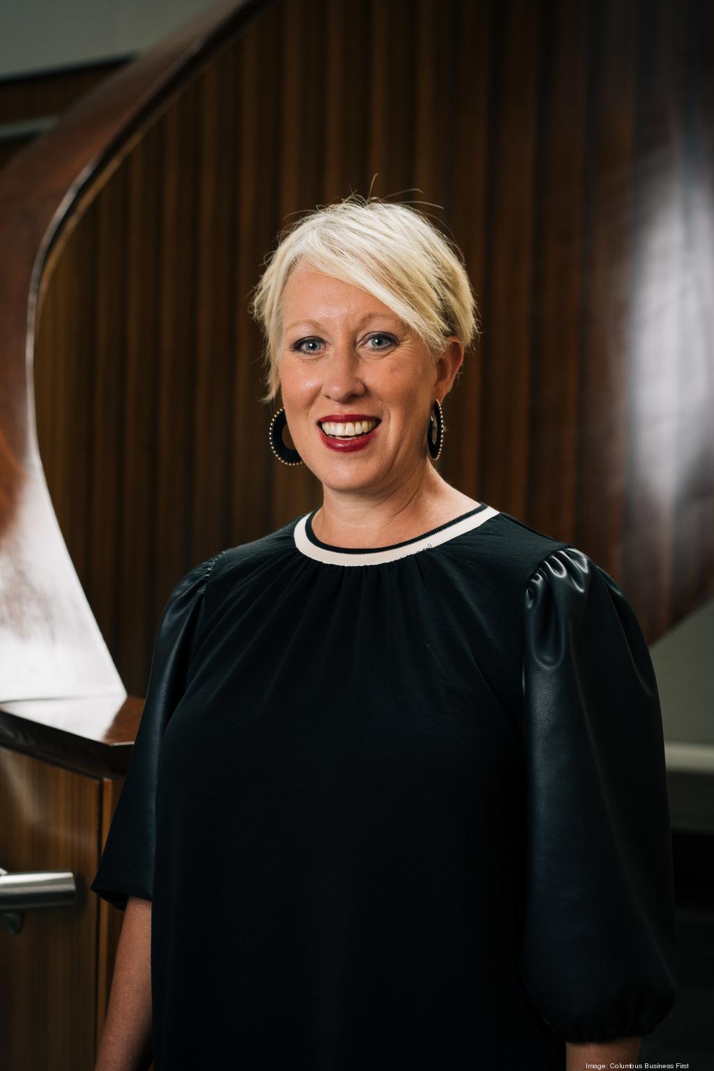 A woman with short blonde hair and hoop earrings, wearing a black dress with puff sleeves, stands in front of a wooden spiral staircase designed by Crimson Design Group.