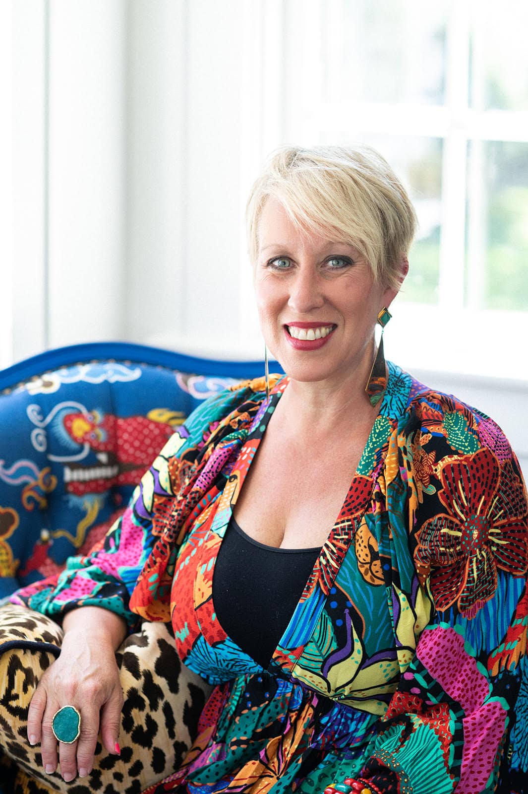 A woman with short blonde hair wearing a colorful, patterned outfit sits on a blue chair with a vibrant animal design by Crimson Design Group, smiling at the camera.