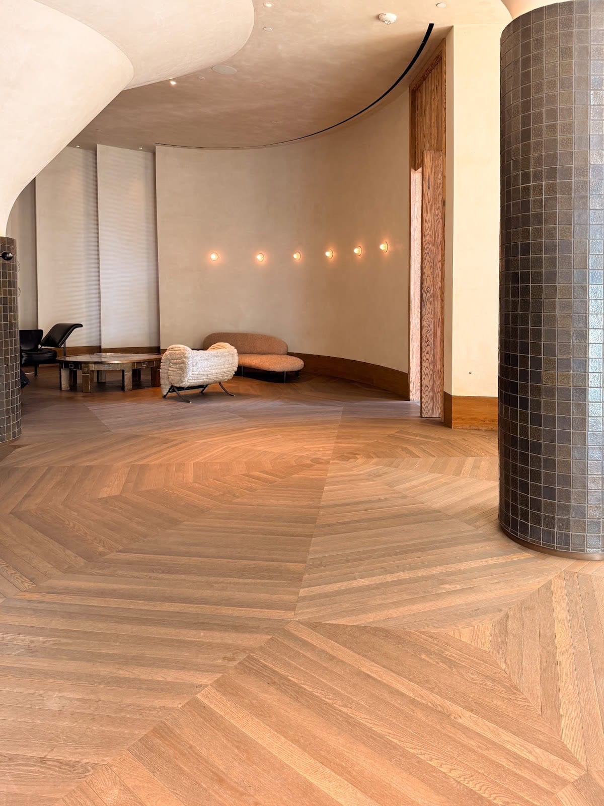 Modern interior with light wood chevron flooring, curved beige walls, tiled column, and minimalist furniture including a round coffee table and two lounge chairs.