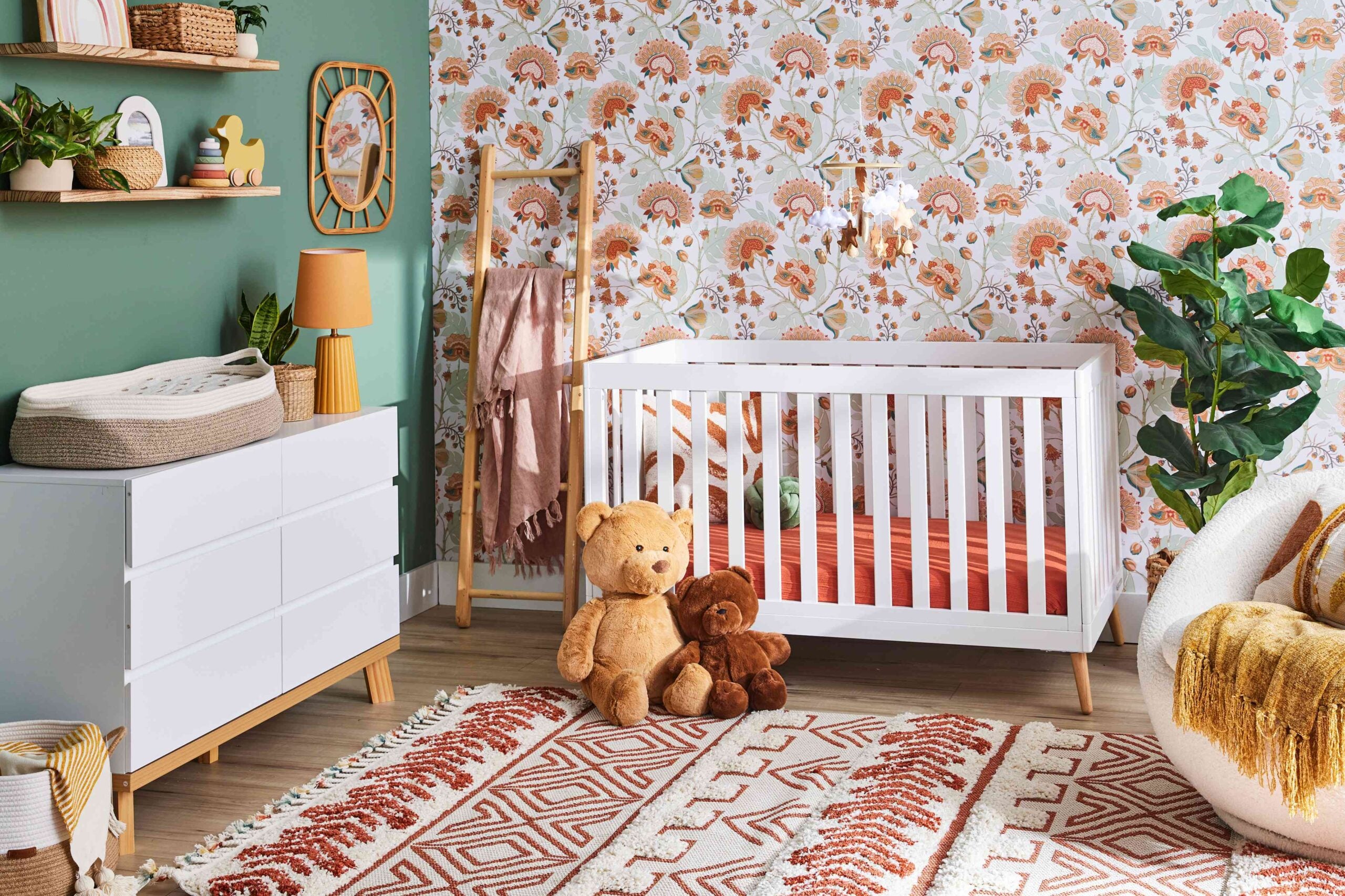 A nursery by Crimson Design Group with a white crib, two stuffed bears on a patterned rug, a dresser with changing pad, leafy plant, and floral wallpaper.