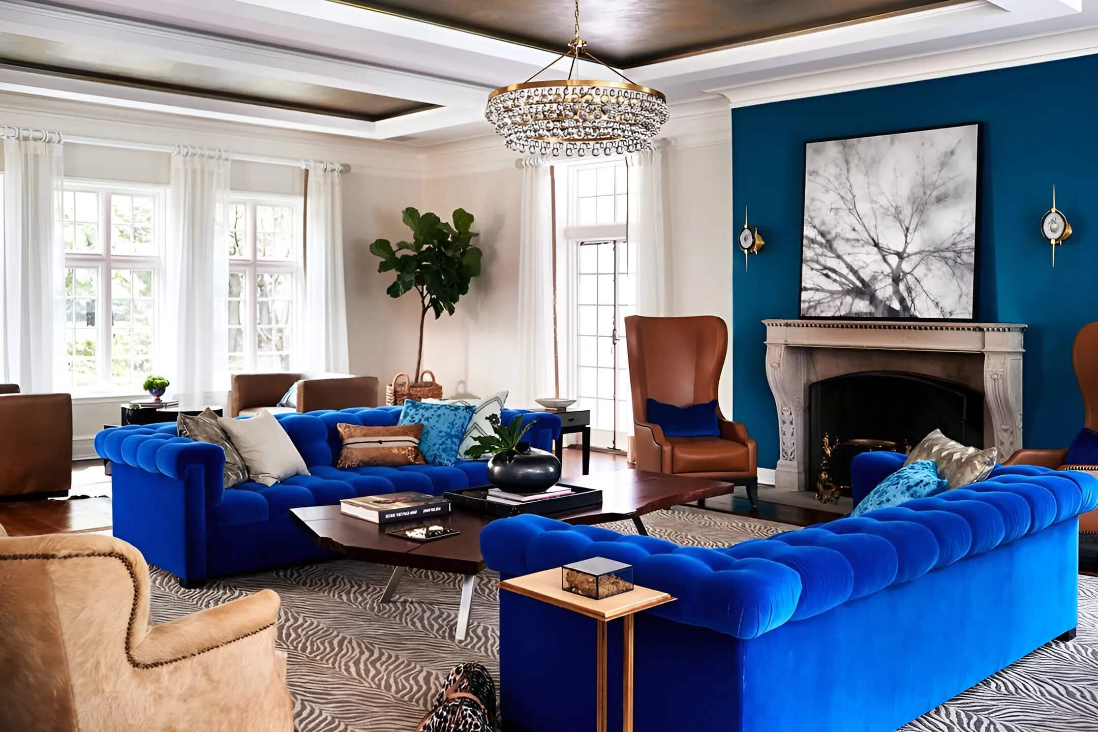 A modern living room with blue velvet sofas, tan armchairs, a fireplace, large windows, a chandelier, and a black-and-white tree artwork above the mantel.