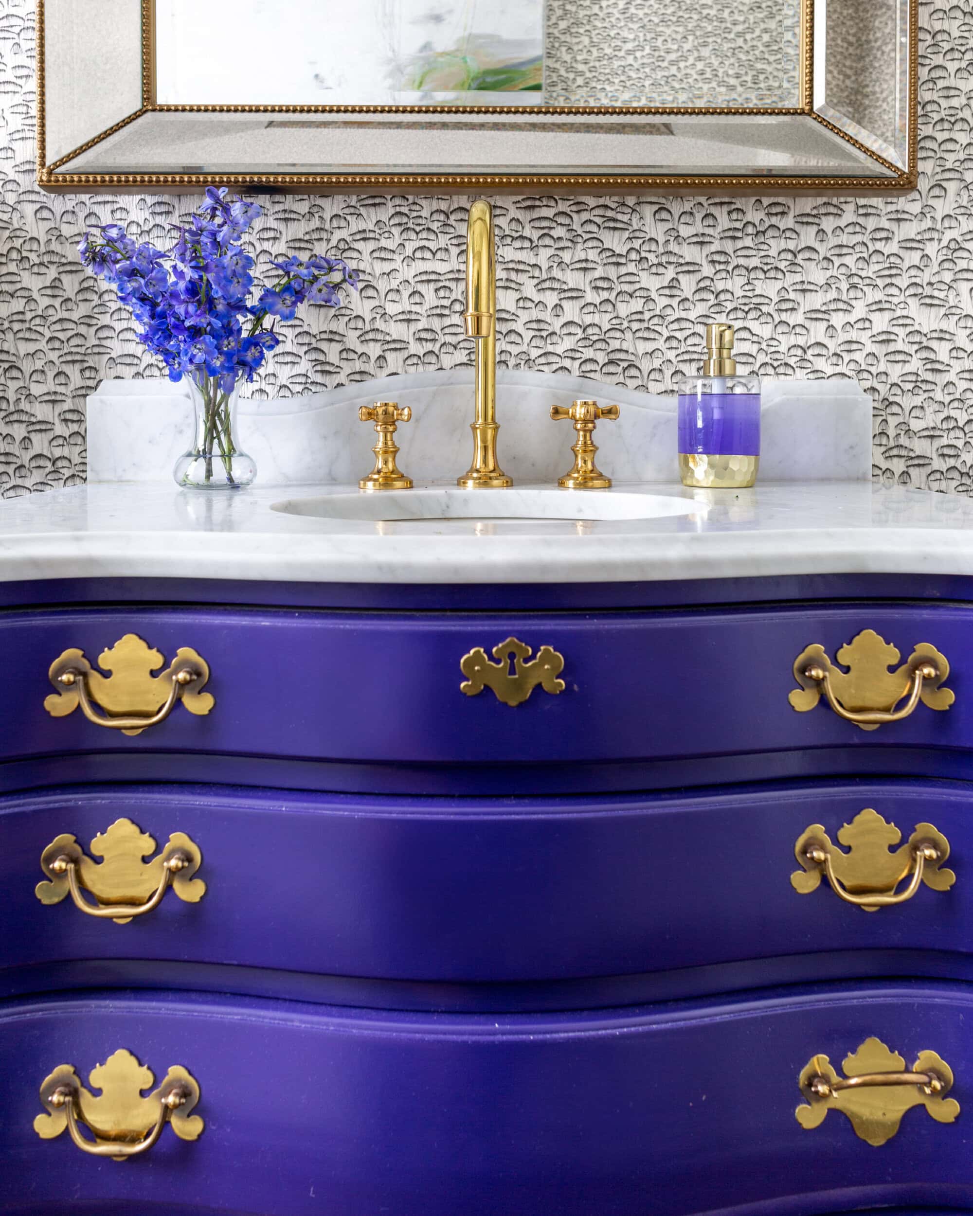A bathroom vanity with a curved, deep purple cabinet, gold hardware, a marble countertop, gold faucet, purple soap dispenser, and a vase of purple flowers.